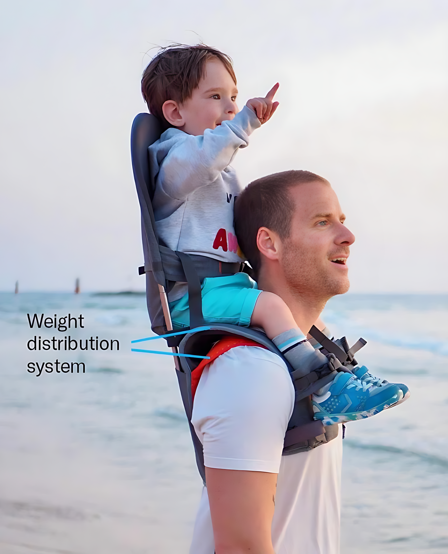 A man carrying a child on his back in a MiniMeis G4 Blue to show the MiniMeis weight distribution system, set against a beach backdrop.
